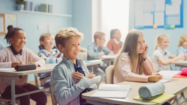 Elementært klasserom av lyssky barn ler sammen etter noe morsomt. De hører på læreren sin som gir lekser. Happy Young Kids i barneskolen Skriver i Øvelseshefter – stockfoto