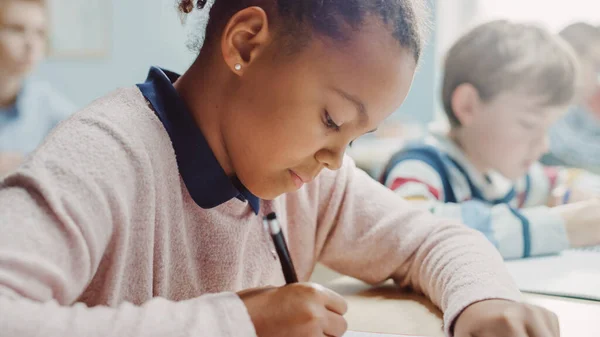 In Elementary School Classroom Girl Writes in Exercise Notebook, Taking Test (en inglés). Aula Junior con Diverso Grupo de Niños Brillantes Trabajando Diligentemente, Aprendiendo. Retrato de vista lateral —  Fotos de Stock