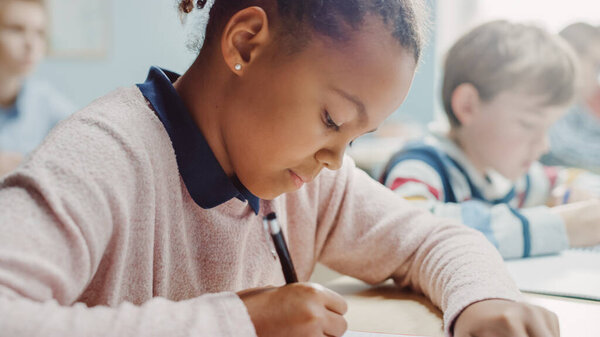 In Elementary School Classroom Girl Writes in Exercise Notebook, Taking Test. Junior Classroom with Diverse Group of Bright Children Working Diligently, Learning. Side View Portrait