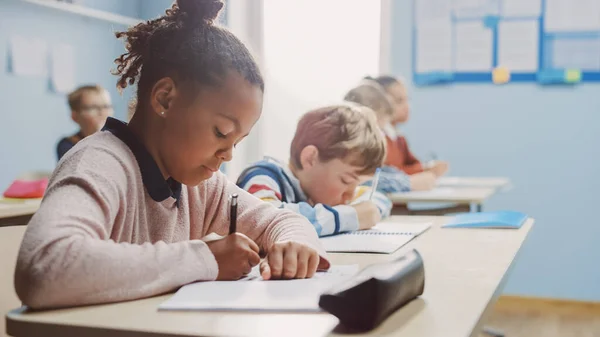 In Elementary School Classroom Brilliant Black Girl Writes in Exercise Notebook, Taking Test. Junior Classroom with Diverse Group of Bright Children Working Diligently and Learning. Side View Portrait — Stock Photo, Image