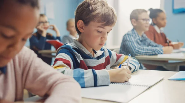 In Elementary School Class: Portrait of Brilliant Caucasian Boy Writes in Exercise Notebook, Taking Test and Writing Exam (en inglés). Grupo Diverso de Niños Brillantes Trabajando Diligentemente y Aprendiendo —  Fotos de Stock