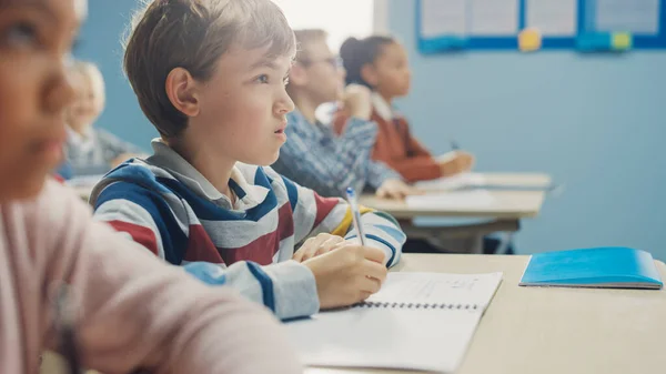 W klasie szkoły podstawowej: Portret genialnego chłopca rasy kaukaskiej pisze w zeszycie z ćwiczeniami, zdając test i egzamin pisemny. Różnorodna grupa jasnych dzieci pilnie pracujących i uczących się — Zdjęcie stockowe
