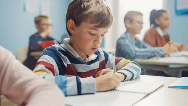 W klasie szkoły podstawowej: Portret genialnego chłopca rasy kaukaskiej pisze w zeszycie z ćwiczeniami, zdając test i egzamin pisemny. Różnorodna grupa jasnych dzieci pilnie pracujących i uczących się — Zdjęcie stockowe