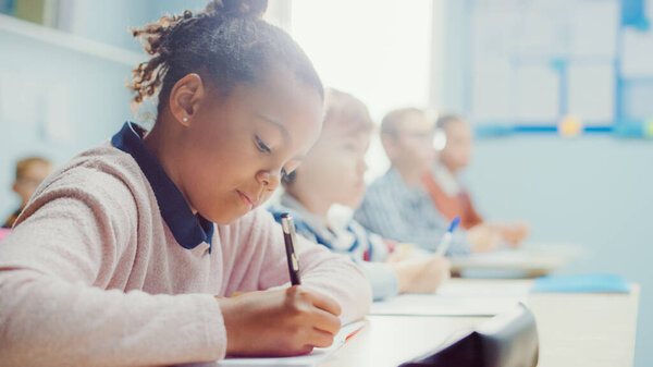 In Elementary School Classroom Black Girl Writes in Exercise Notebook, Taking Test. Junior Classroom with Diverse Group of Bright Children Working Diligently, Learning. Low Angle Side View Portrait