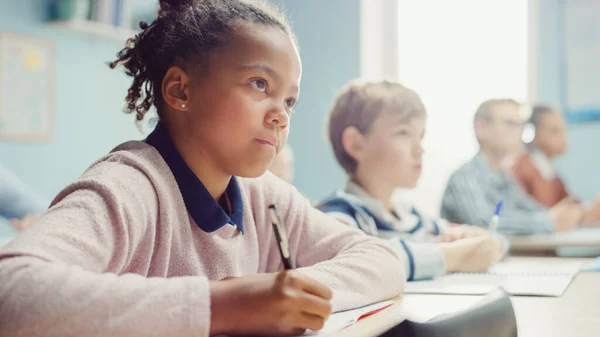 Az általános iskolában a fekete lány ír a gyakorló jegyzetfüzetbe, vizsgázik. Junior tanterem a változatos csoport Bright Children dolgozik szorgalmasan, tanulás. Alacsony szögű Side View Portré — Stock Fotó