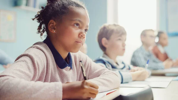 En el aula de la escuela primaria, Black Girl escribe en el cuaderno de ejercicios, tomando prueba. Aula Junior con Diverso Grupo de Niños Brillantes Trabajando Diligentemente, Aprendiendo. Retrato de vista lateral de ángulo bajo — Foto de Stock