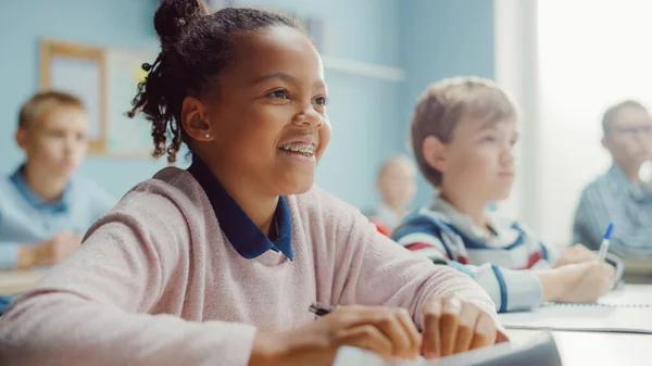 Dans la classe de l'école primaire : Portrait d'une fille noire brillante avec des sourires de bretelles, écrit dans le carnet d'exercices. Salle de classe junior avec un groupe diversifié d'enfants apprenant de nouvelles choses — Photo