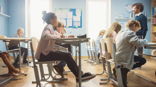 En la clase de la escuela primaria: El maestro entusiasta camina entre filas de niños brillantes y diversos, dicta la lección. Grupo de niños multiétnicos inteligentes que escriben en cuadernos de ejercicios . —  Fotos de Stock