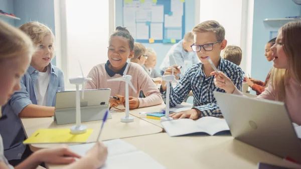 En la Escuela Primaria: Clase de Niños Pequeños Inteligentes Trabajan en Equipo Usando Computadoras Tablet para Programar Turbinas Eólicas. Aula con niños Aprendiendo sobre formas ecológicas de energía renovable —  Fotos de Stock