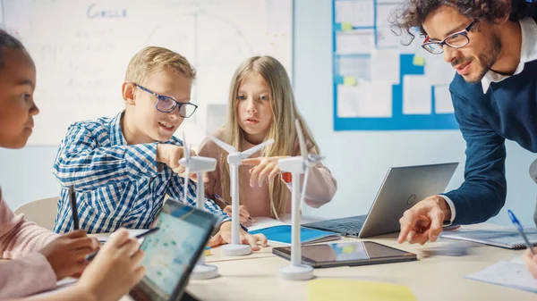 Aula de la escuela primaria: Profesor entusiasta sosteniendo la tableta La computadora explica a un niño pequeño brillante cómo funcionan las turbinas de viento. Niños aprendiendo sobre las formas ecológicas de energía renovable — Foto de Stock