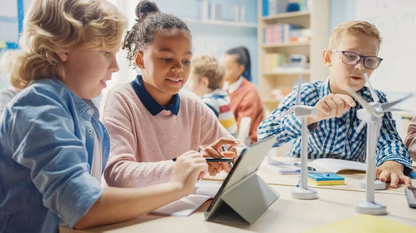 En la Escuela Primaria: Clase de Niños Pequeños Inteligentes Trabajan en Equipo Usando Computadoras Tablet para Programar Turbinas Eólicas. Aula con niños Aprendiendo sobre formas ecológicas de energía renovable —  Fotos de Stock