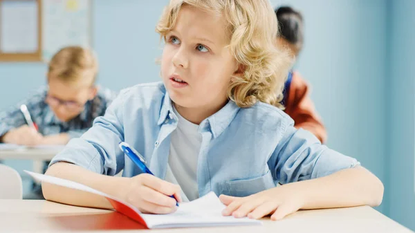 I grundskolan klass: Porträtt av en lysande och söt vit pojke skriver i övning anteckningsbok. Junior klassrum med olika grupper av ljusa barn arbetar flitigt, lära sig nya saker — Stockfoto