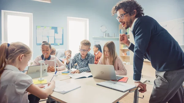 En la Escuela Primaria: Clase de Niños Pequeños Inteligentes Trabajan en Equipo Usando Computadoras Tablet para Programar Turbinas Eólicas. Aula con niños Aprendiendo sobre formas ecológicas de energía renovable — Foto de Stock