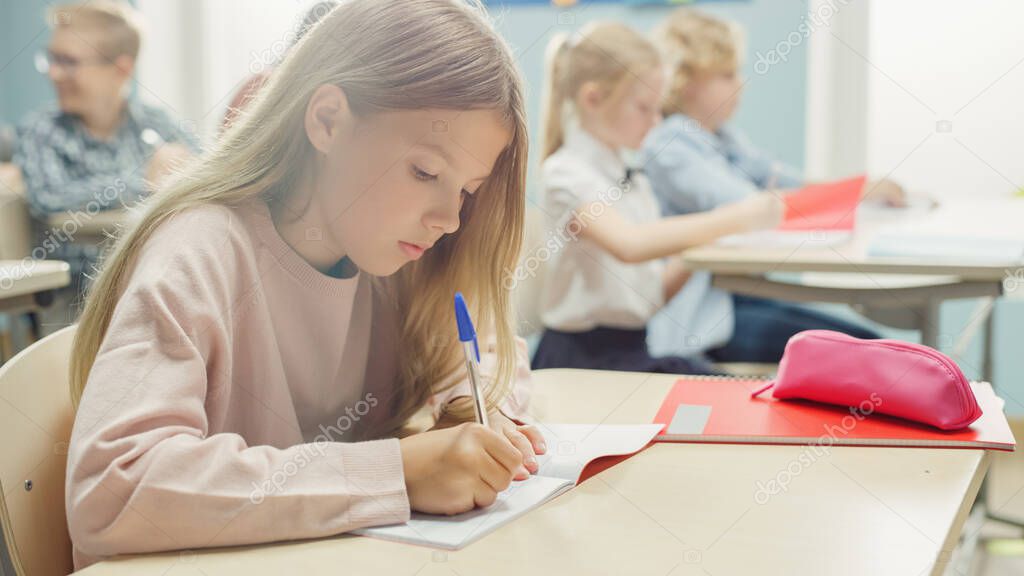 In Elementary School Classroom Caucasian Girl Writes in Exercise Notebook, Taking Test and Writing Exam. Junior Classroom with Diverse Group of Children Working Diligently and Learning New Stuff
