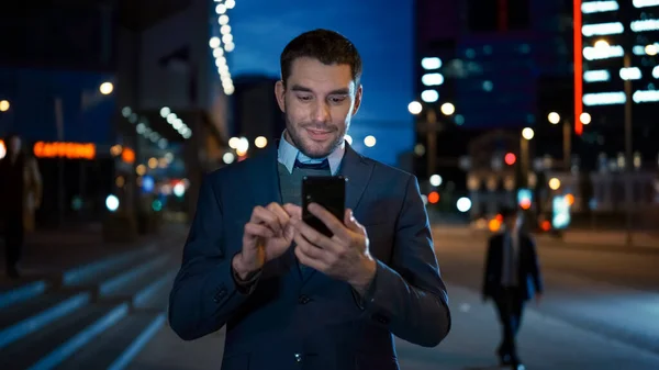 Портрет кавказского бизнесмена в костюме использует смартфон на Темной улице в вечернее время. Он выглядит уверенным и успешным. Атмосферные огни города на заднем плане . — стоковое фото