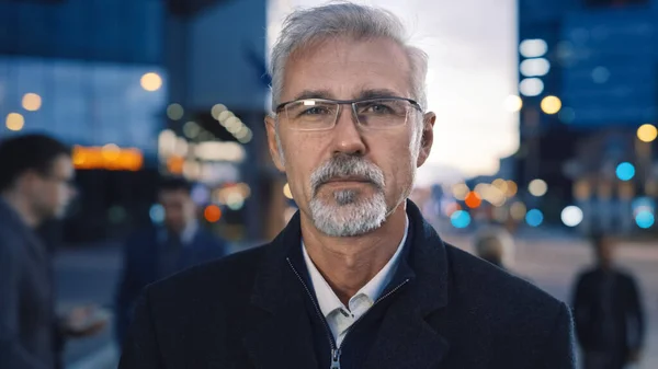 Portret poważnego starszego biznesmena w płaszczu stojącego na ulicy z pieszymi. Ma okulary z brodą i okularami. Wieczór z oświetleniem miejskim w tle. — Zdjęcie stockowe