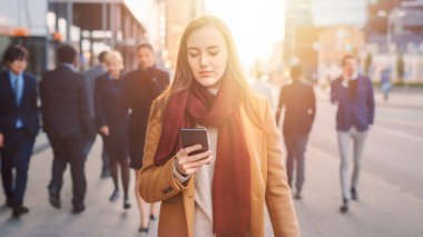Genç tesadüfen giyinmiş kadın caddede akıllı bir telefon kullanıyor. İş adamları ve ofis yöneticileri işe giderken yolda yürürler. Telefonunu kontrol ederken kendinden emin görünüyor. Ilık Güneş parıltısıyla çekilen.