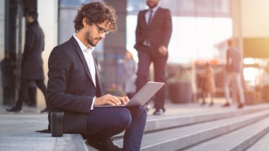 Takım Elbiseli Yakışıklı İş Adamı İş Merkezi 'nin yanındaki basamaklarda oturuyor ve şehirdeki bir sokakta dizüstü bilgisayarda çalışıyor. Ofisteki insanlar işe yürür. Ilık Güneş parıltısıyla çekilen.