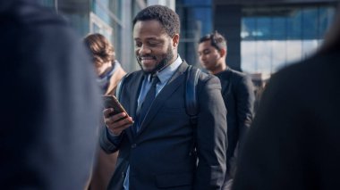 Sokakta yayalarla dikilen takım elbiseli Afrikalı Amerikalı bir iş adamının portresi. - Akıllı telefon kullanıyor. - Evet. Başarılı birine benziyor. Diğer İnsanlar İşe Gidiyor.