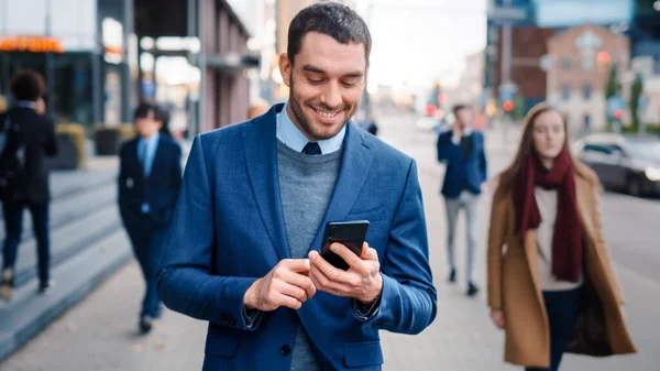 Blanke zakenman in een pak gebruikt een smartphone in een straat in het centrum. Andere kantoormensen lopen voorbij. Hij lacht en ziet er succesvol uit. Hij surft op het web op zijn apparaat. — Stockfoto