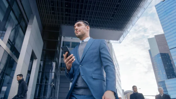 Kavkazský podnikatel v obleku používá chytrý telefon na ulici v centru. Jiní lidé z kanceláře procházejí minulostí. Usmívá se a vypadá, že uspěl. Prohlížení webu na svém zařízení. — Stock fotografie