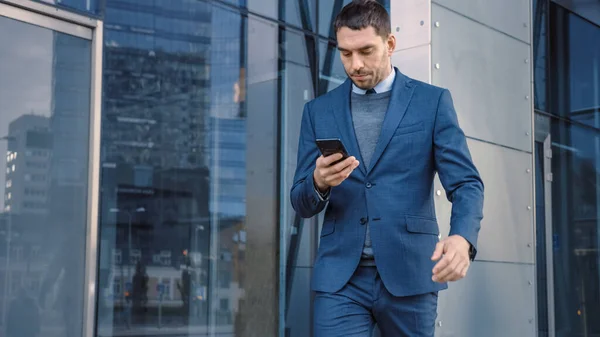 Vit affärsman i kostym använder en smartphone på en gata i centrum. Andra kontor Människor går förbi. Han är självsäker och ser framgångsrik ut. Han bläddrar på webben på sin enhet. — Stockfoto