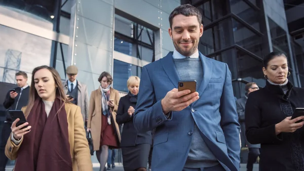 Toimistopäälliköt ja liikemiehet kävelevät modernin lasitoimistorakennuksen edessä ja käyttävät älypuhelimiaan. Ihmiset ovat pukeutuneet älykkäästi ja näyttävät onnistuneilta. He ovat käyttäneet laitteitaan. . — kuvapankkivalokuva