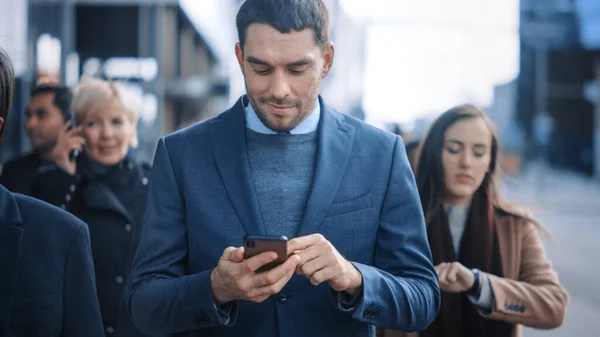 Blanke zakenman in een pak gebruikt een smartphone in een straat in het centrum. Andere kantoormensen pendelen in een menigte. Hij is zelfverzekerd en lijkt succesvol. Hij surft op het web op zijn apparaat. — Stockfoto