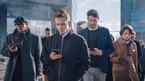 Kantoormanagers en zakenmensen lopen voor een modern glazen kantoorgebouw en gebruiken hun smartphone. Mensen zijn slim gekleed en zien er succesvol uit. Ze zijn bezet door hun apparaten. — Stockfoto