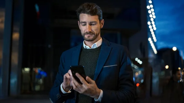 Ψηλός Καυκάσιος Επιχειρηματίας με στολή χρησιμοποιεί ένα Smartphone στη Σκοτεινή Οδό το βράδυ. Περιπλανιέται ενώ φαίνεται σίγουρος και επιτυχημένος. Ατμοσφαιρική Αστική Πόλη Φώτα στο παρασκήνιο. — Φωτογραφία Αρχείου