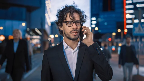 Nuori tyylikäs liikemies, jolla on kiharat hiukset, puhuu puhelimessa kävellessään kaupungissa. Hänellä on silmälasit päässään. Toimistoväki kävelee ohi. Sen ilta ilmakehän kaupunkivalojen kanssa taustalla. — kuvapankkivalokuva