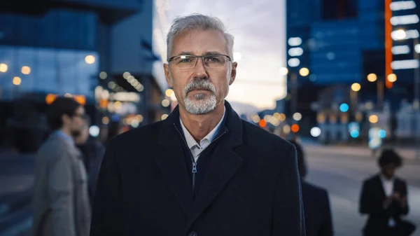 Portrett av en seriøs senior forretningsmann i en frakk stående på en gate med fotgjengere. Han har skjegg og briller på seg. Kveld med atmosfærisk urbane lys i bakgrunnen. – stockfoto