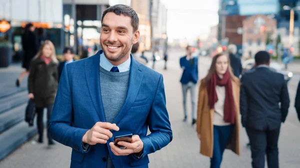 Vit affärsman i kostym använder en smartphone på en gata i centrum. Andra kontor Människor går förbi. Han ler och ser framgångsrik ut. Han surfar på nätet på sin enhet. Skjuten med varm sol fackla. — Stockfoto
