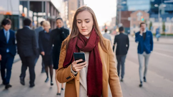 Νεαρή έξυπνη γυναίκα ντυμένη τυχαία χρησιμοποιεί ένα Smartphone σε ένα δρόμο. Επιχειρηματίες και διευθυντές γραφείων περπατούν στο πέρασμα στο δρόμο τους για την εργασία. Φαίνεται σίγουρη ενώ τσεκάρει το κινητό της και περπατάει.. — Φωτογραφία Αρχείου