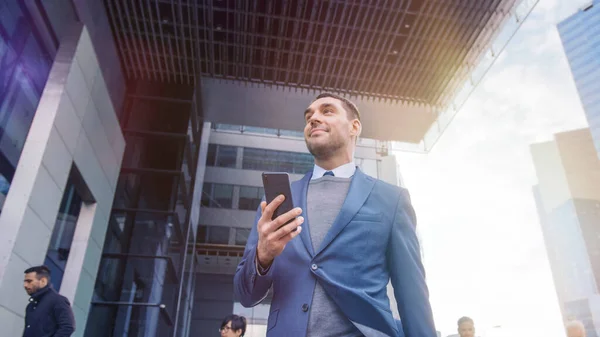 En hvit forretningsmann i dress bruker en smarttelefon på en gate i sentrum. Andre kontorfolk går forbi. Han smiler og ser vellykket ut. Han Browsing the Web på sin Device. – stockfoto