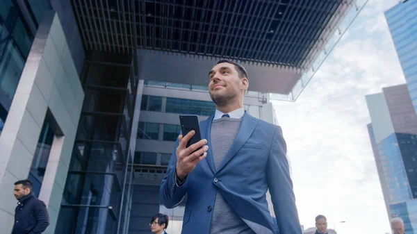 Valkoihoinen liikemies puvussa käyttää älypuhelinta kadulla keskustassa. Muut ihmiset kävelevät menneisyydessä. Hän hymyilee ja näyttää onnistuneelta. Hän selailee verkkoa laitteellaan.. — kuvapankkivalokuva