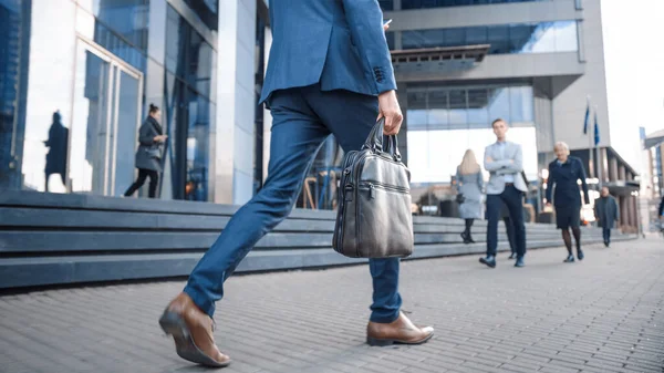 Primer plano de la pierna tiro de un hombre de negocios en un traje de viaje a la oficina a pie. Lleva una maleta de cuero. Otros gerentes y gente de negocios caminan cerca. Día nublado en una calle del centro . —  Fotos de Stock