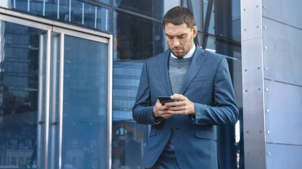 En hvit forretningsmann i dress bruker en smarttelefon på en gate i sentrum. Andre kontorfolk går forbi. Han er selvsikker og fremgangsrik. Han Browsing the Web på sin Device. – stockfoto