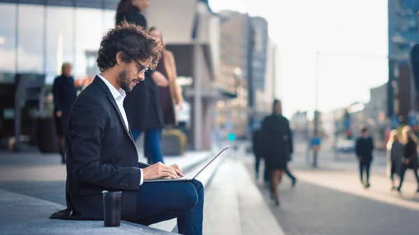 Snygg affärsman i kostym sitter på trappan bredvid Business Center och arbetar på en bärbar dator på en gata i en stad. Kontor Människor går förbi för att arbeta. — Stockfoto