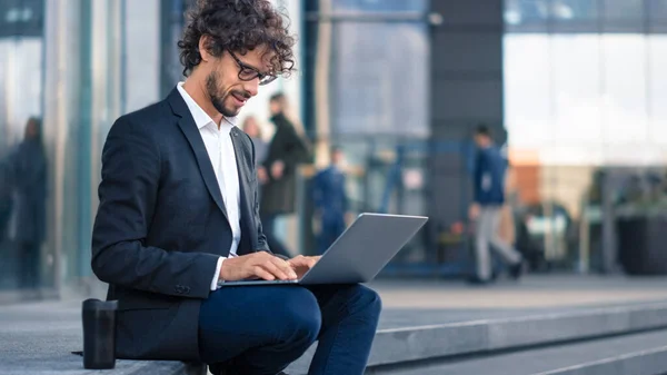 Handsome Businessman in a Suit zit op Steps naast Business Center en werkt op een laptop in een straat in een stad. Kantoormensen lopen langs om te werken. — Stockfoto