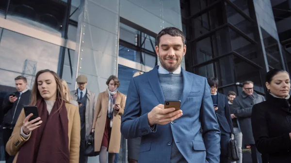 Kontorschefer och affärsmän går framför en modern glaskontorsbyggnad och använder sina smartphones. Folk är snyggt klädda och ser framgångsrika ut. De är ockuperade av sina enheter. — Stockfoto