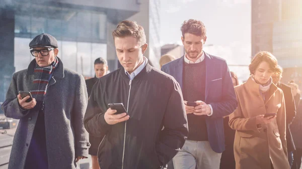 Kantoormanagers en zakenmensen lopen voor het moderne glazen kantoorgebouw en gebruiken hun smartphones. Mensen zijn slim gekleed. Ze worden bewoond door hun apparaten. Schot met warme zonnevlam — Stockfoto