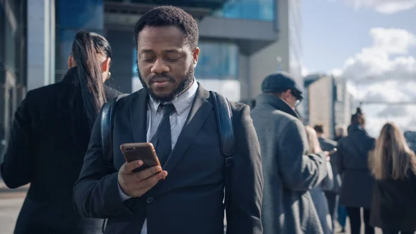 Portret van een Afro-Amerikaanse zakenman in een pak staand in een straat met voetgangers. Hij gebruikt een smartphone. Hij ziet er succesvol uit. Andere mensen gaan aan het werk. — Stockfoto