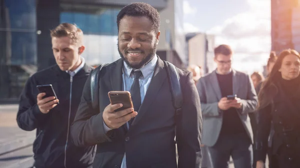Portret van een lachende Afro-Amerikaanse zakenman in een pak staand in een straat met voetgangers. Hij gebruikt een smartphone. Hij ziet er succesvol uit. Andere mensen gaan aan het werk. — Stockfoto