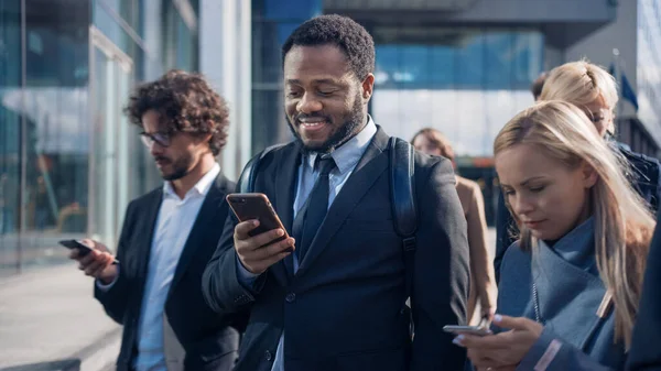 Portrett av en afrikansk-amerikansk forretningsmann i dress stående på en gate med fotgjengere. Han bruker en smarttelefon. Han ser vellykket ut. Andre går på jobb. – stockfoto