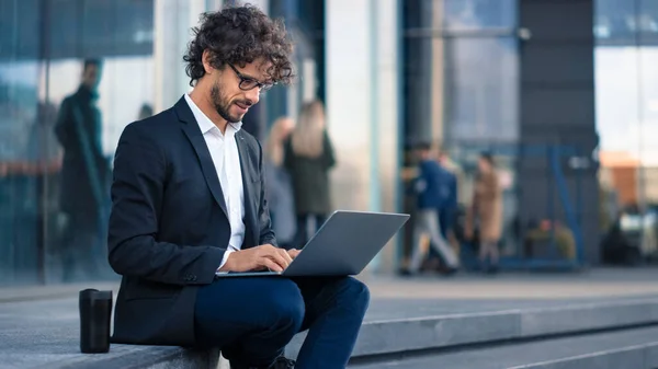 Bonito homem de negócios em um terno está sentado em degraus ao lado do centro de negócios e trabalhando em um laptop em uma rua em uma cidade. Pessoas de escritório caminham para o trabalho . — Fotografia de Stock