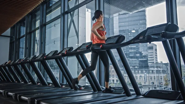Beautiful Athletic Sports Woman Running on a Treadmill. Energetic Fit Female Athlete Training in the Gym Alone. Urban Business District Window View. Side Back View