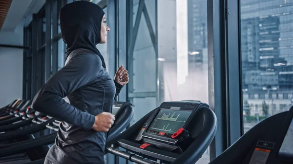 Athletic Muslim Sports Woman Wearing Hijab and Sportswear Running on a Treadmill (en inglés). Entrenamiento de atleta femenina Energetic Fit en el gimnasio solo. Vista de la ventana del distrito de negocios urbano. Retrato de vista lateral —  Fotos de Stock