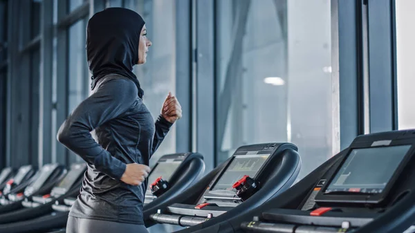 Athletic Muslim Sports Woman Wearing Hijab and Sportswear Running on Treadmill. Energetic Fit Female Athlete Training in Gym Alone. Urban Business District Window View. Side View Portrait