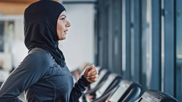 Femeia sportivă atletică musulmană care poartă hijab și îmbrăcăminte sportivă alergând pe banda de alergare. Energetic Fit feminin atlet de formare în sala de sport singur. Urban Business District Window View. Portret vedere laterală — Fotografie, imagine de stoc